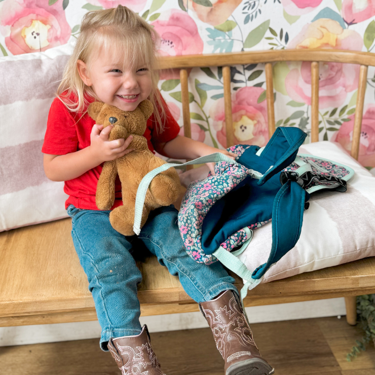 A little, blond girl with cowboy boots smiling and hugging a teddy bear.