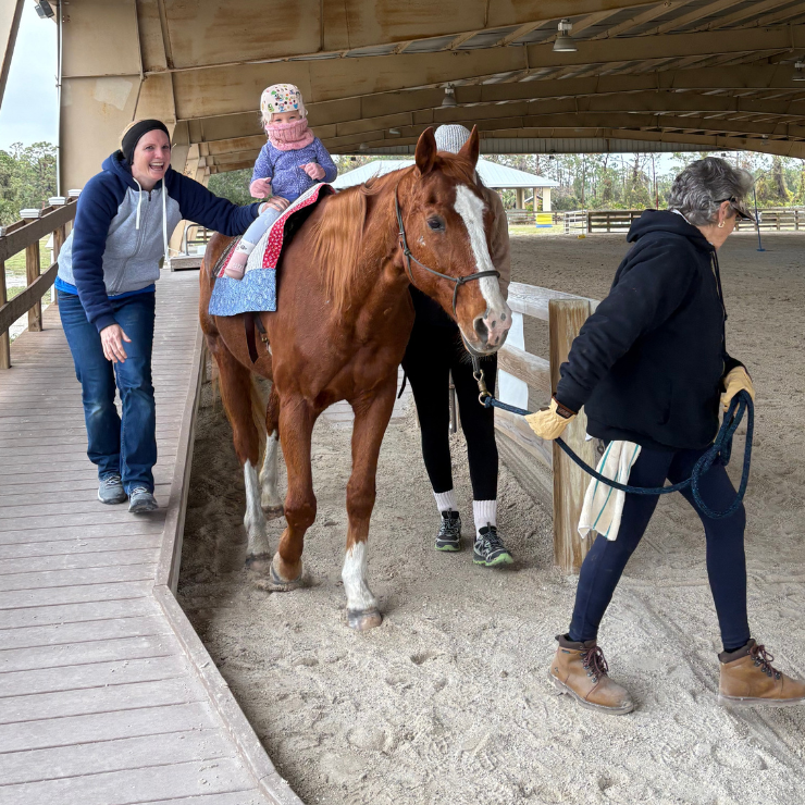 A little girl riding a horse surrounded by adults at a therapy session.