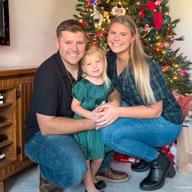 A man, woman and little girl smiling in front of a Christmas tree.