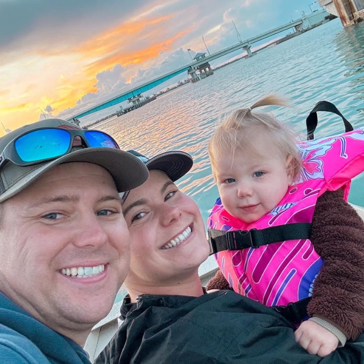 A man, woman and little girl smiling with a sunset and water in the background. 