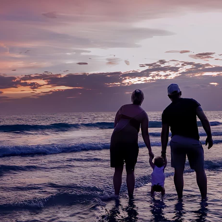A woman and man leaning over to hold the hands of their toddler on the beach with the sun setting and their backs to the camera. 