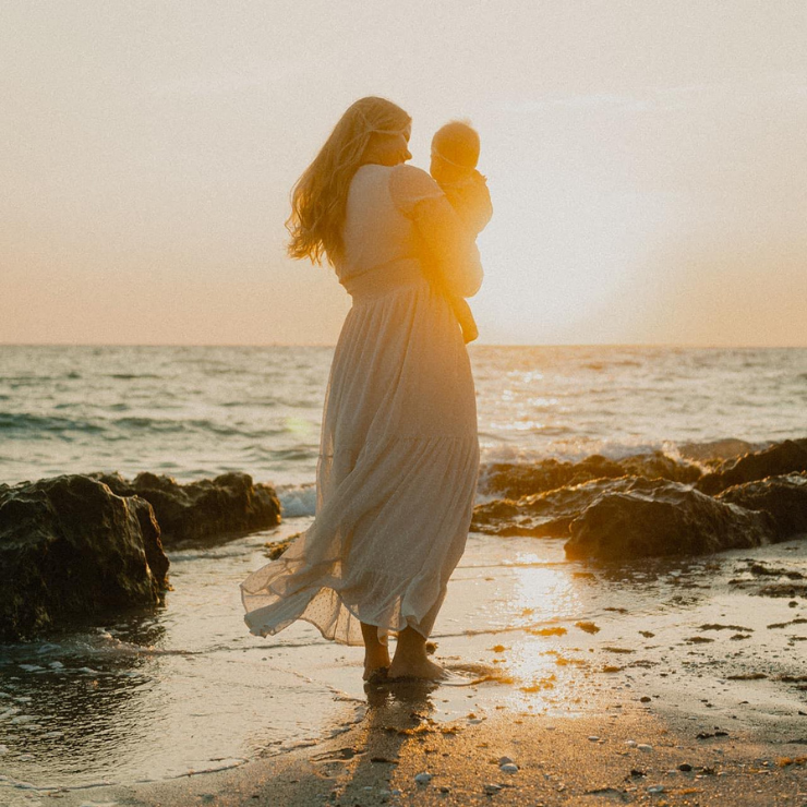 A woman in a white dress holding a baby with their back to the camera and the sun shining behind them on the beach. 