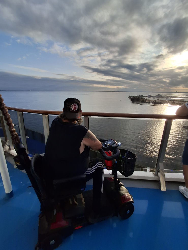 Person on a mobility scooter on a blue deck facing the ocean at sunset or sunrise with a cloudy sky.
