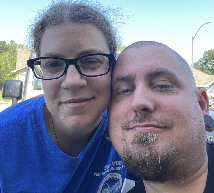 Two people smiling outdoors with a clear blue sky in the background. One is wearing glasses and a blue shirt; the other has a short haircut.