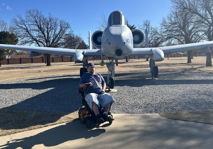 Man in a motorized wheelchair in front of a plane with a blue sky in the background