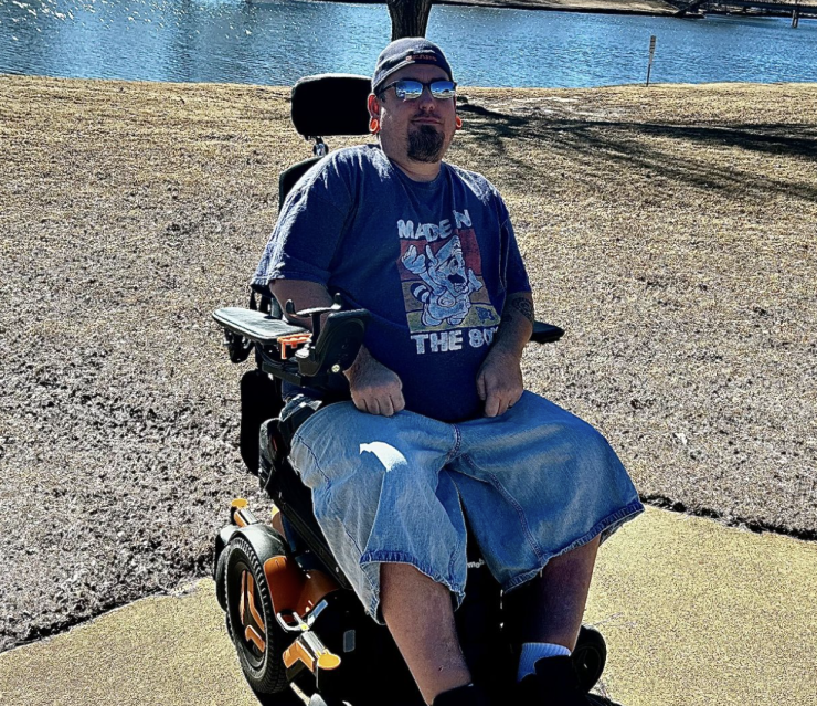 Man in a wheelchair by a lake with trees and a bright sky in the background.