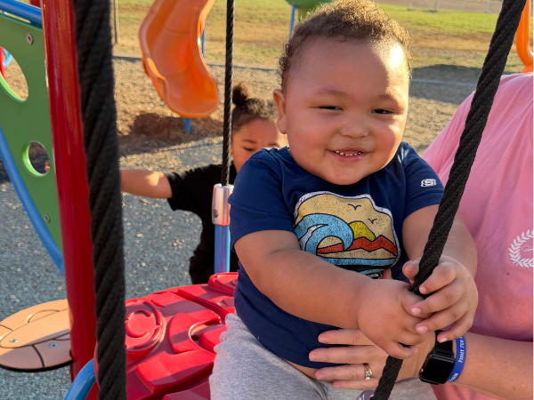 Smiling child holding the ropes of a playground swing while supported by an adult.