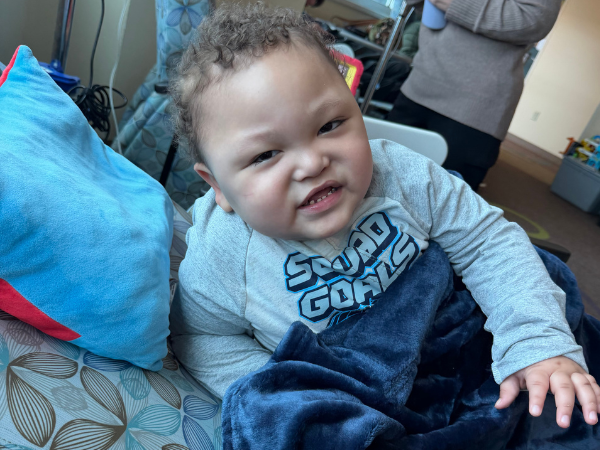 Smiling young child seated with a blue blanket beside a bright blue pillow, wearing a shirt that says “SUPER COOL.”