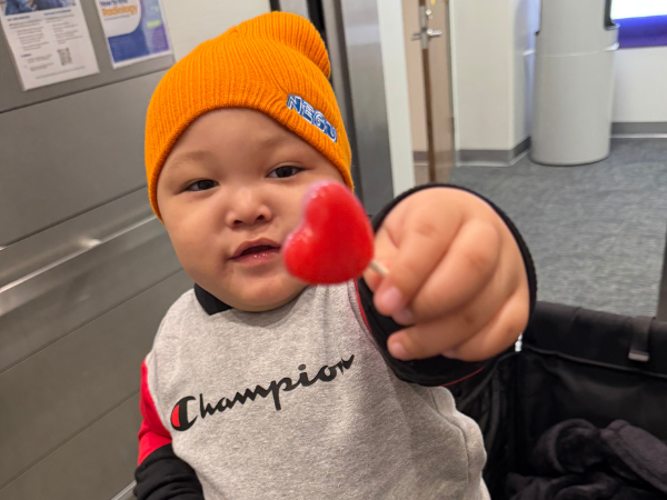 hild in an orange beanie holds a red heart-shaped lollipop toward the camera while seated in a stroller.