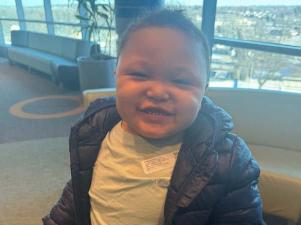 Smiling child in a dark quilted jacket sitting near large windows overlooking a city.