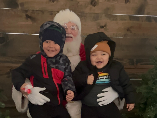 Two children in winter clothing pose with Santa Claus in front of a rustic wooden wall.