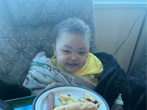 Smiling toddler wrapped in blankets sits behind a plate of a sandwich, sausage, and crinkle-cut fries.